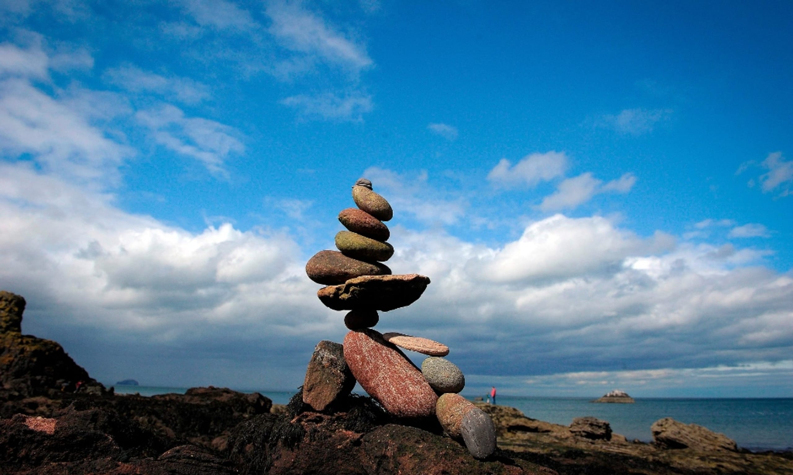 Balanced rock cairn standing on a coastal shoreline under a bright blue sky with clouds